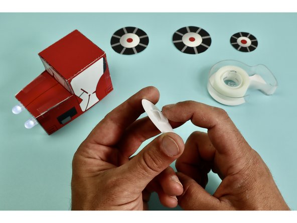 Using clear tape make a loop of tape on the back of the two large wheels. These wheels get attached to the back of the tractor where it says &quot;clear tape.&quot;