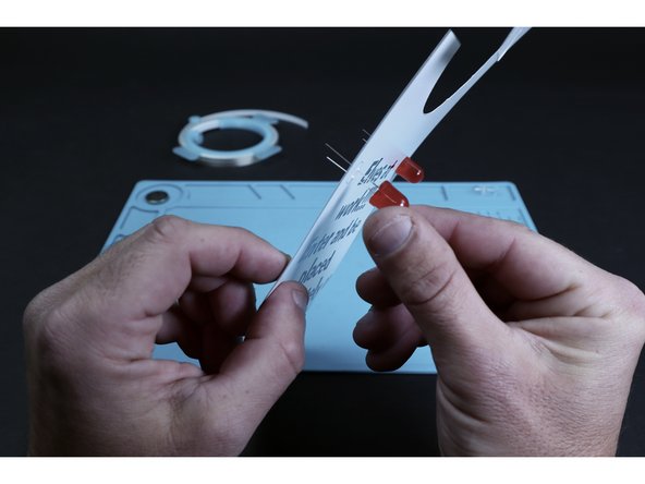 Each of the pairs of holes you just poked is for a single LED. From the front, thread the legs of each of your two LEDs into one of those pairs of small holes. Take care to insert the long LED legs into the upper holes and the shorter LED legs into the bottom holes.