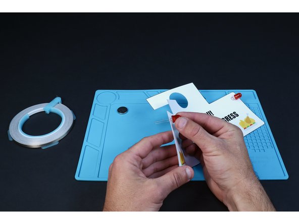 Each of the pairs of holes you just poked is for a single LED. From the front, thread the legs of each of your two LEDs into one of those pairs of small holes. Take care to insert the long LED legs into the upper holes and the shorter LED legs into the bottom holes.