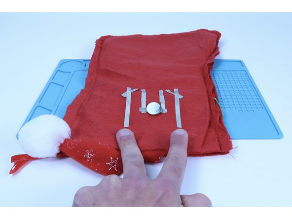 Recall that the two outer legs are the longer (+) legs. Cut two 3&quot; pieces of Maker Tape. Peel and stick one to each of those two outer (+) legs and then TO the stocking (half on the leg/half on the felt).  The ends of these paths should extend beyond the battery as shown.