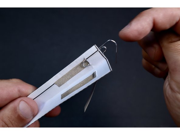 Seat the paperclip within the folds as shown so that the small curly bend is threaded through the slit and the part you bent outward is oriented toward the center fold.