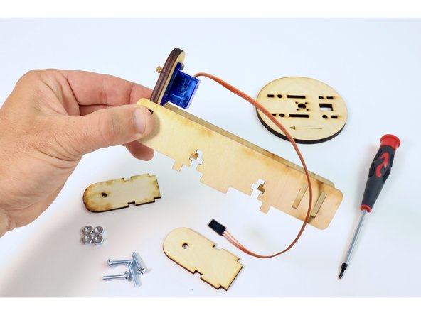 Seat the tabs of the servo mount in the single slots of the two long wooden pieces as shown. Pinch and hold them in place making sure the servo axle is facing outward as shown with the servo itself on the opposite side of the nut/screw slots of those long wooden pieces.