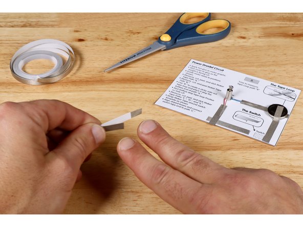 Wrap Maker Tape around the small switch template as shown. This step requires a single piece of Maker Tape that starts at the fold line on one side the template, wraps around to cover the entirety of the opposite side and hangs off that end by 1/4&quot;.