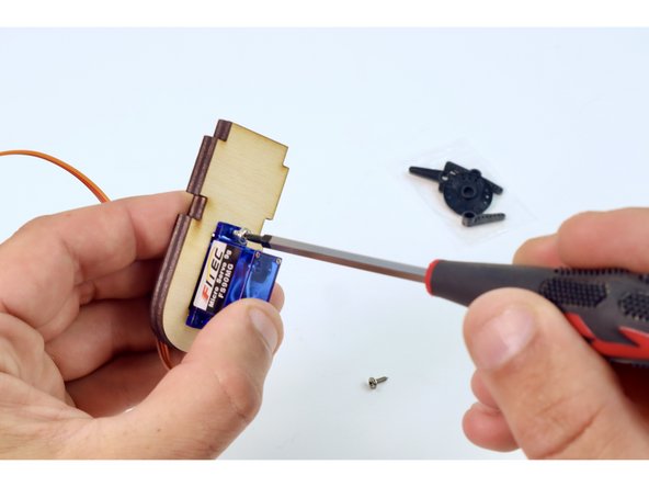 Using the wooden servo plate shown, and the two matching small screws found in the last servo bag, mount the remaining servo  using your Phillips head screwdriver.