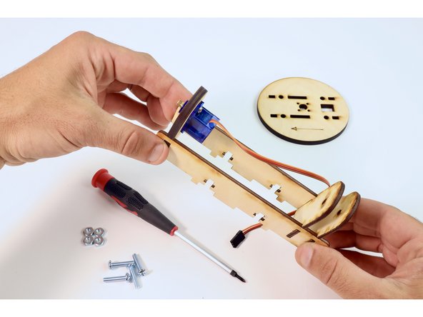 Insert the remaining two matched wooden pieces into the slots at the opposite end of the longer pieces. Make sure their curved ends match the orientation of the curved end of the servo plate. Pinch and hold together.