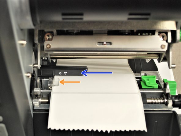 Feed the labels through/under the black ribbon sensor (blue arrow in the picture) and clear label guide (orange arrow in picture).
