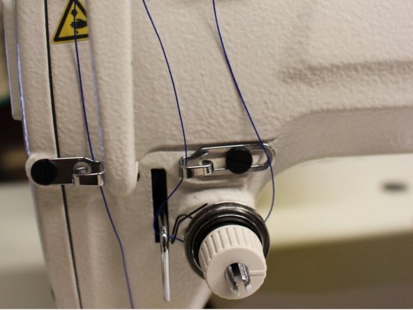 At the bottom front left, there is a knob and a few silver hooks surrounding it. Wrap the thread from right to left in a clockwise motion so the thread wraps the bottom of the knob.