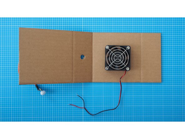 Use the double sided tape to stick the fan to the backside of the 3rd cardboard segment using the tape.  Make sure the fan is centered and that the scored lines of the cardboard are facing down as shown here.