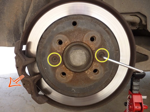 Desserrer le frein à main et tourner le disque de frein afin de trouver le boulon de réglage via le trou rendu disponible suite à la dépose de l'obturateur (repère bleu).