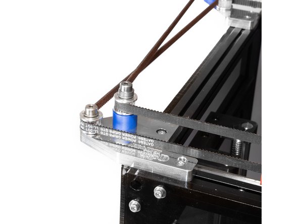 The belt crosses the left side of the printer, to the toothed idler on the tall stack of the rear idler mount.
