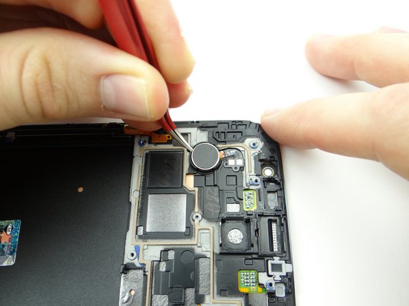 Wedge the curved-tip tweezers in the closed position under the vibrator precisely where pictured.  Lift the vibrator, but stop when you reach the vibrator cable.