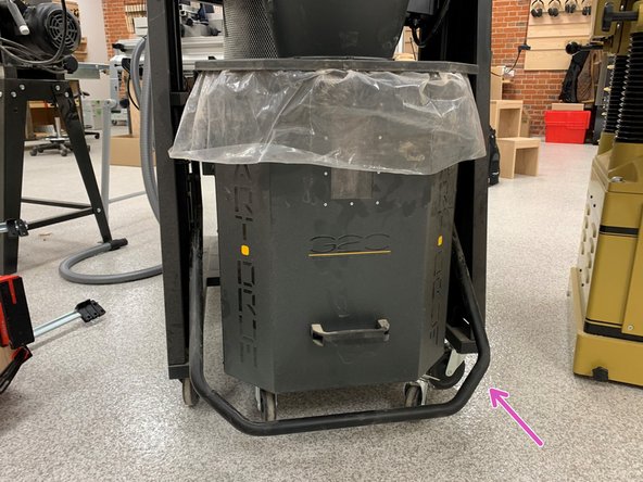 Undo the metal latch holding the band around the top of the waste bin. Tip over and empty into sawdust barrel.